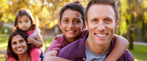A family of four enjoys time together in park, as the son hangs off the father's back and the daughter snuggles with the mother
